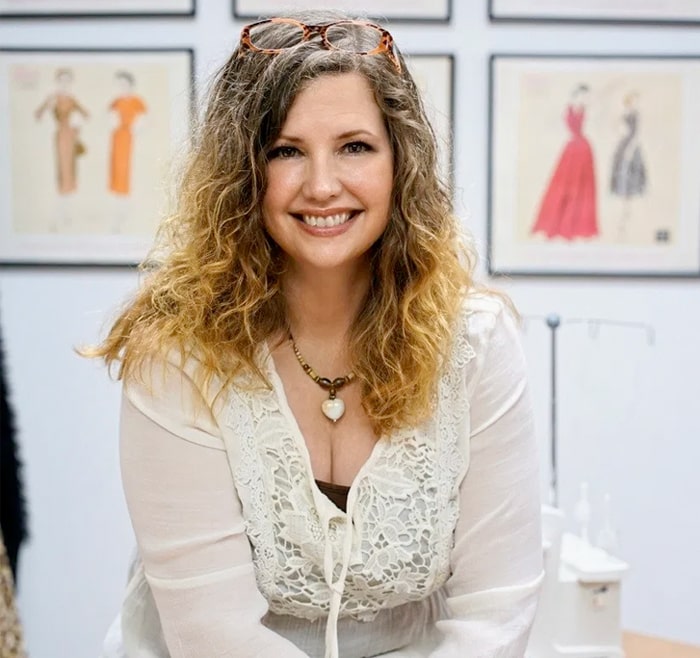 A smiling woman with curly hair wearing a light blouse and sunglasses on her head, seated in front of fashion illustrations inspired by sewing classes in Minneapolis.